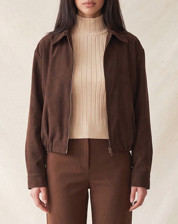 A model wearing a dark brown suede jacket over a beige ribbed turtleneck sweater and matching brown trousers, standing against a neutral textured beige backdrop.