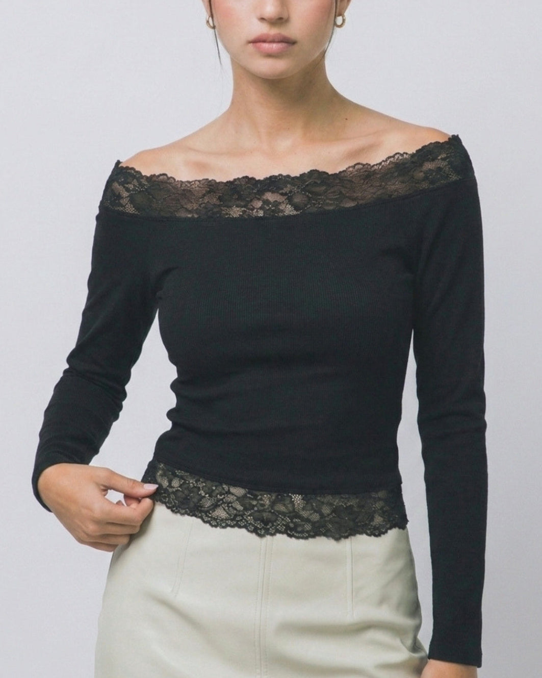 A woman modeling a black off-the-shoulder long-sleeve top with delicate black lace trim across the neckline and waist, paired with a light beige skirt, against a plain light gray background.