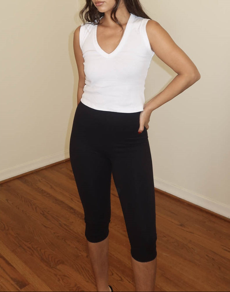 A woman modeling a white sleeveless V-neck top and black capri leggings in an indoor room with beige walls and a wood floor.