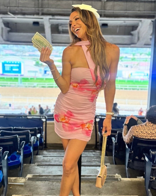 A woman stands in a racetrack stadium wearing a light pink halter mini dress with a pink floral print, holding a stack of cash in one hand and a beige handbag in the other. The background shows empty stadium seats and a racetrack.