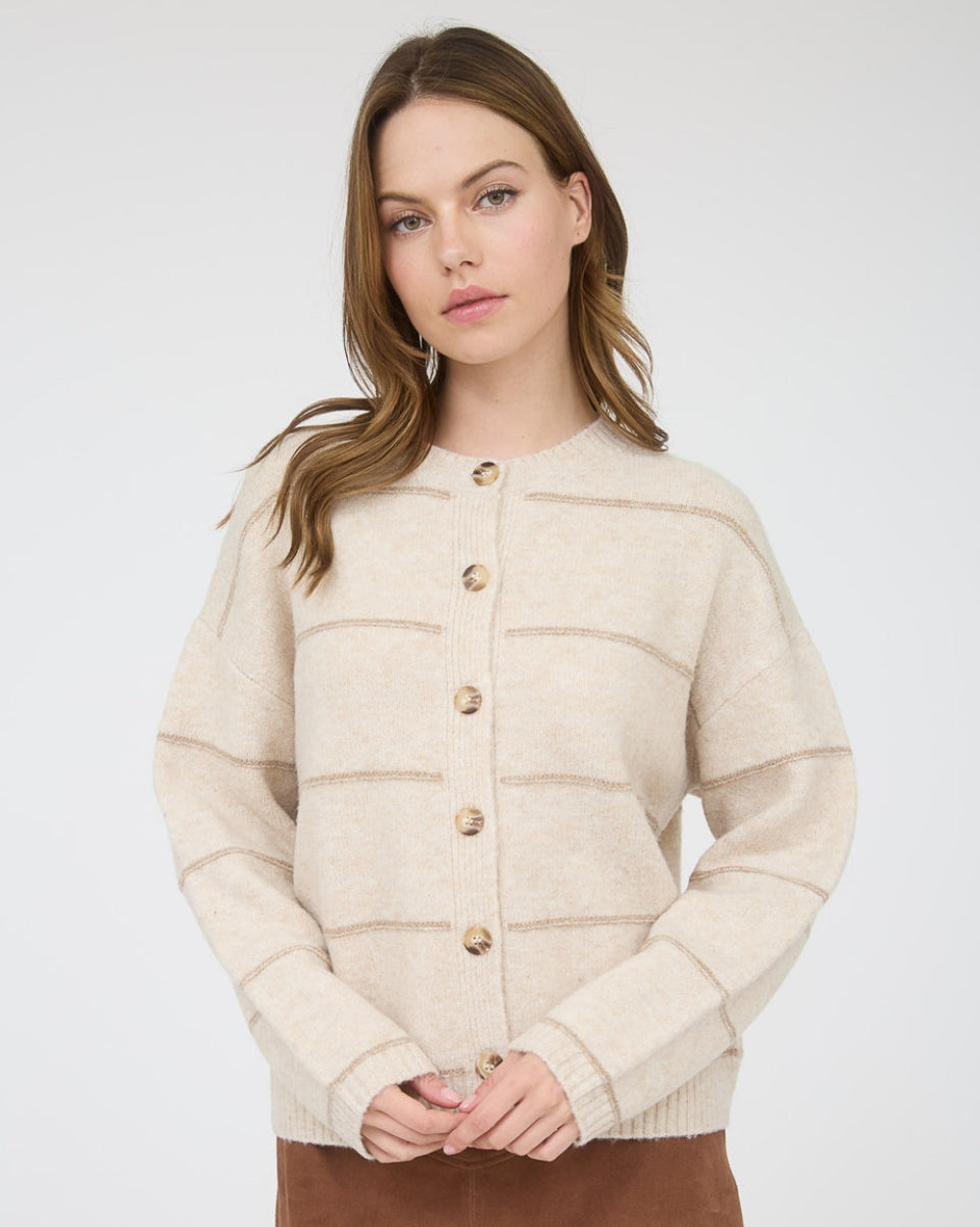A woman modeling a cream-colored knit cardigan with horizontal textured stripes and gold buttons, paired with a brown mini skirt, against a light gray studio background.