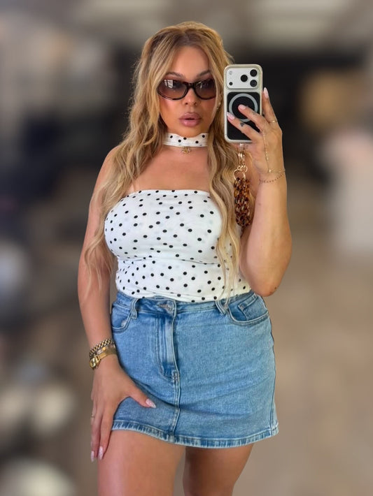 A woman taking a mirror selfie wearing a white strapless top with black polka dots, a white choker necklace, and a light blue denim mini skirt.