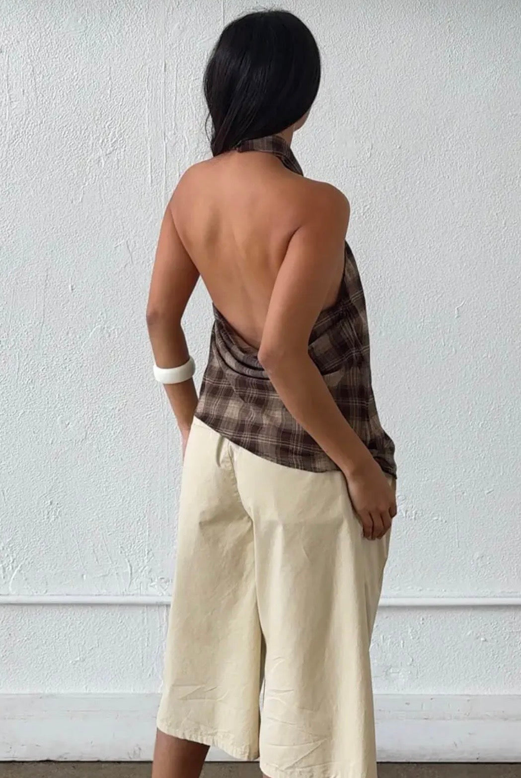 A woman shown from behind wearing a brown and beige plaid halter top with an open back and cream-colored wide-leg shorts, standing against a white textured wall.