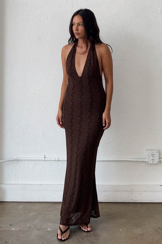 A woman modeling a dark brown lace halter maxi dress with a deep V-neckline, standing against a white textured wall.