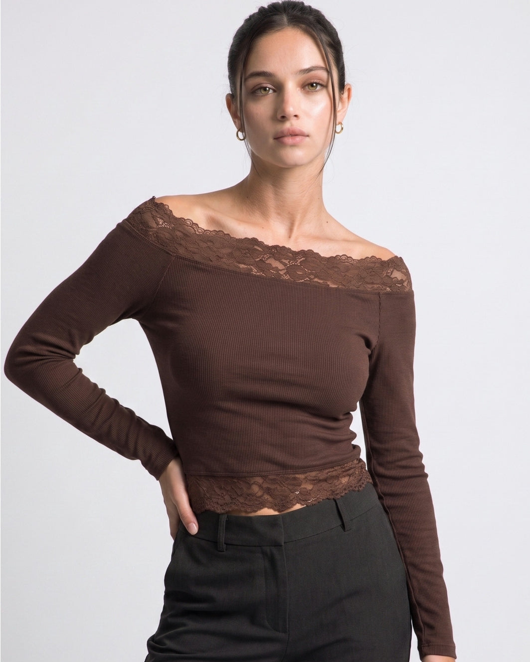 A woman modeling a brown off-the-shoulder long-sleeve top with lace trim, paired with black pants, against a light gray studio background.