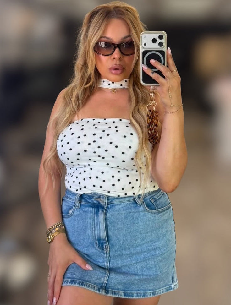 A woman taking a mirror selfie wearing a white strapless top with black polka dots, a white choker necklace, and a light blue denim mini skirt.