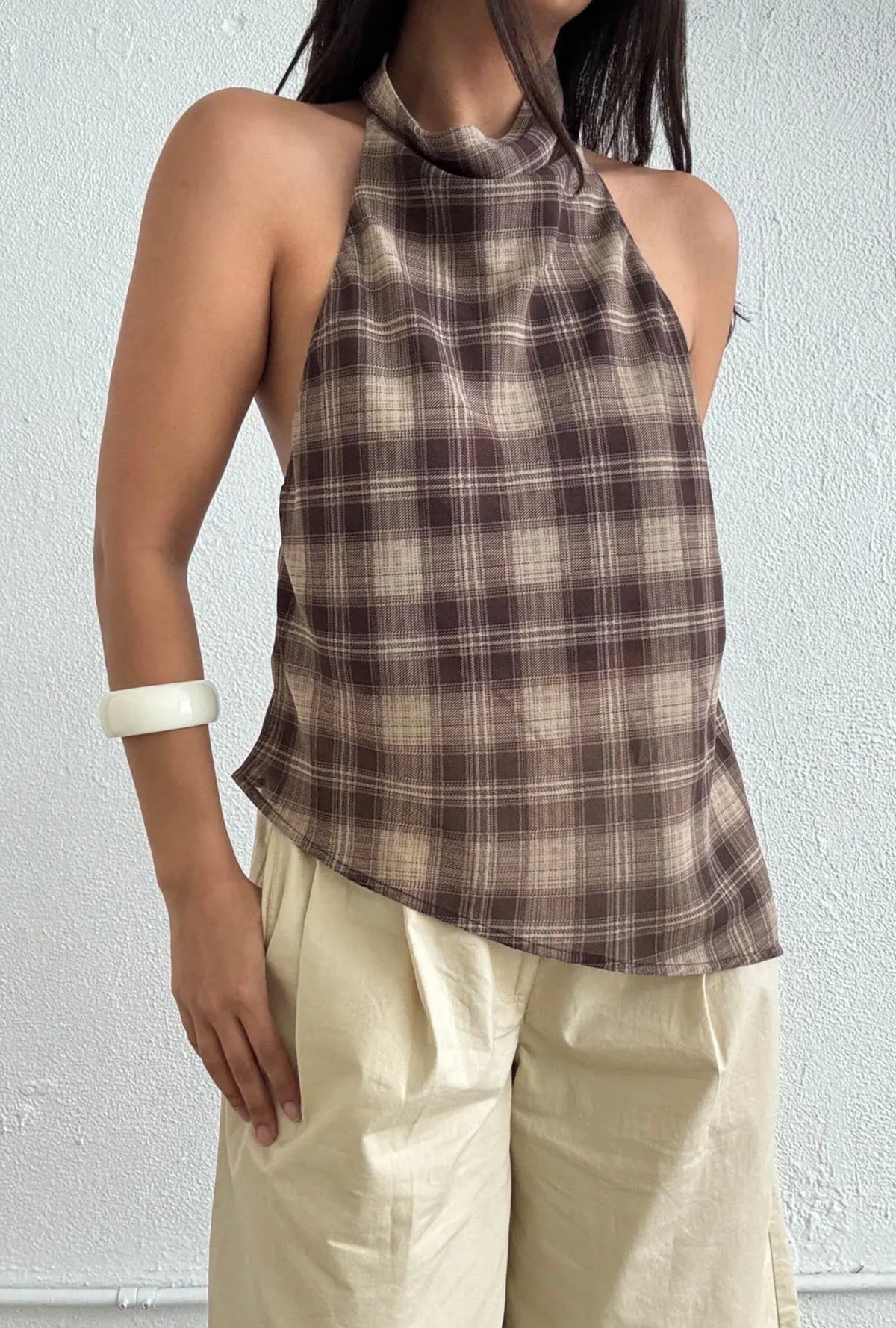 A woman wearing a brown and beige plaid halter top paired with cream-colored pants, standing against a light textured wall.