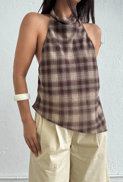 A woman wearing a brown and beige plaid halter top paired with cream-colored pants, standing against a light textured wall.